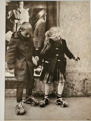 Kids unsteady on skates in parisian street with tonton flingueur in shop window behind by Michael Joseph
